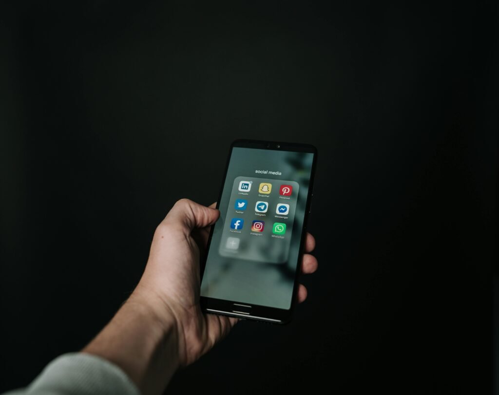 Best Side Hustles to Make Money in 2025 Close-up of a hand holding a smartphone displaying various social media app icons on a dark background.